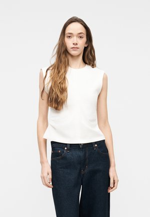 Młoda kobieta z długimi brązowymi włosami, ubrana w białą bluzkę bez rękawów i ciemnoniebieskie jeansy z wysokim stanem, na jednolitym tle.