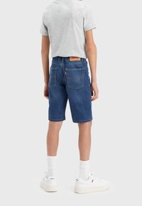 Pantalones cortos de mezclilla en un tono azul oscuro, con bolsillos traseros con costuras amarillas y una etiqueta, combinados con una camiseta gris y zapatillas blancas.