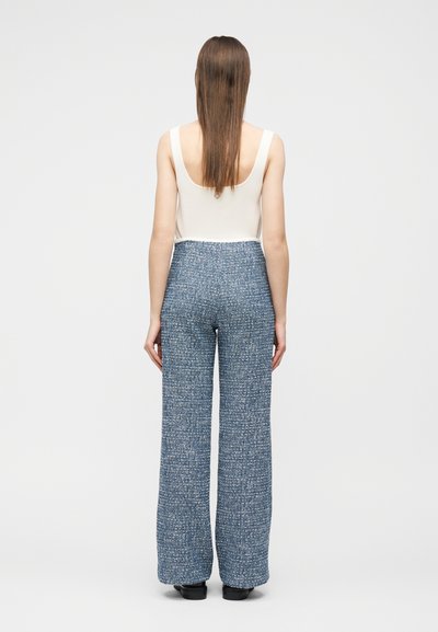 Γυναίκα με μακριά ίσια μαλλιά που φοράει λευκό αμάνικο τοπ και ψηλόμεσα μπλε υφασμάτινα παντελόνια με φαρδιά μπατζάκια, στέκεται με την πλάτη προς τα εμπρός.