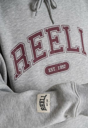Gray sweatshirt with maroon "REEL" print and "EST. 1997" text. Features a tag on the sleeve and a hood with drawstrings. Soft fabric texture.