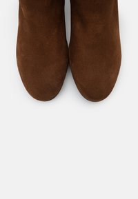 Botas de ante marrón hasta el tobillo con punta redondeada y textura suave, que presentan un diseño elegante adecuado para diversas ocasiones.