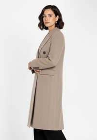 Beige dubbelknäppt rock med ett texturerat tyg, som har en sjalkrage, två framfickor och svarta knappar. Skräddarsydd silhuett.