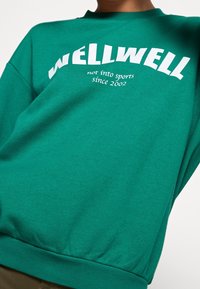 Grön bomullströja med rundad hals, ribbad mudd och texten "WELLWELL inte intresserad av sport sedan 2002" i fet vit stil.