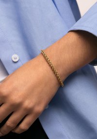 Pulsera trenzada de tono dorado llevada en una muñeca, que presenta una textura suave y un brillo sutil sobre un fondo de camisa azul claro.