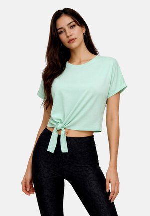 Young woman with long dark hair wears mint green short sleeve crop top tied at the waist and high-waisted black patterned leggings.
