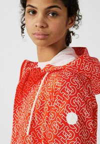 Rode jas met wit logopatroon, voorzien van een capuchon en contrasterende mesh voering. Inclusief witte trekkingkoordjes en een gestructureerd label op de mouw.