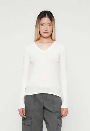 Young woman with straight ombre hair wearing a white V-neck long-sleeve shirt and gray cargo pants against a plain background.