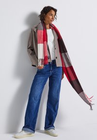 Veste en mélange de laine grise, t-shirt blanc, jeans larges bleus et écharpe à motif à carreaux rouges avec des franges. Des baskets blanches complètent la tenue.