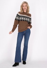 Maglione a maglia marrone con motivi geometrici neri, bianchi e grigi, abbinato a jeans a zampa d'elefante blu e stivaletti neri.