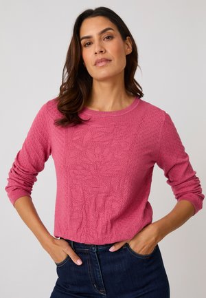 Vrouw draagt een gestructureerde roze trui met lange mouwen en bloemendessin, blauwe spijkerbroek en handen in de zakken tegen een neutrale achtergrond.