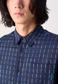 Camisa de botones azul marino con rayas verticales, un bolsillo en el pecho y detalles de costura verde. Material de algodón con un acabado texturizado.