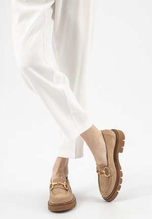 Mocassini in camoscio beige con suole spesse marroni e dettagli in metallo dorato a forma di morsetto, indossati con pantaloni bianchi a sigaretta.