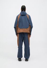 Waterdichte jas en broek set in marineblauw en bruin. Beschikt over een capuchon, een oversized pasvorm en contrasterende kleurvlakken voor een modern ontwerp.