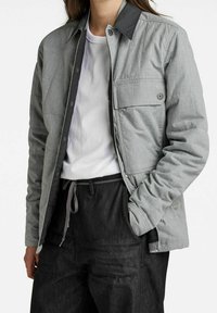 Grau gesteppten Jacke mit einem schwarzen Kragen, ausgestattet mit einer Brusttasche und Druckknöpfen, kombiniert mit dunklen Hosen und einem weißen Hemd.