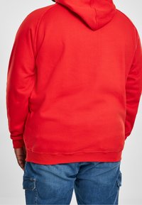 Man die een felrode hoodie en blauwe denim cargobroek draagt, staand met zijn rug naar de kijker, tegen een effen achtergrond.