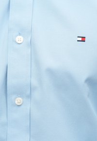 Chemise bleu clair à boutons, fabriquée dans un tissu lisse, avec des boutons blancs et un petit logo brodé aux rayures rouge, blanc et bleu.