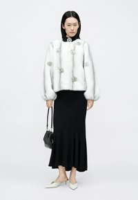 Chaqueta de piel sintética blanca con adornos redondos, combinada con una falda negra estilo sirena. La modelo sostiene un bolso negro texturizado y lleva zapatos blancos puntiagudos.