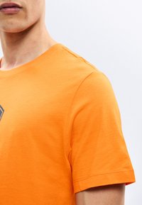 Κοντινό πλάνο ενός ατόμου που φοράει μια φωτεινή πορτοκαλί κοντομάνικη μπλούζα με μερικό σκοτεινό τύπωμα στο στήθος πάνω σε απλό φόντο.