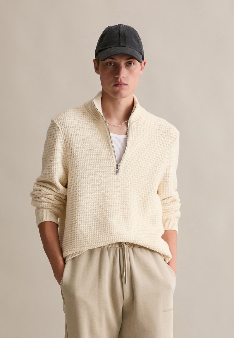 Cremefarbener, strukturierten Pullover mit einem Viertelreißverschluss-Kragen, kombiniert mit beigen Kordelzughosen und einer schwarzen Mütze.