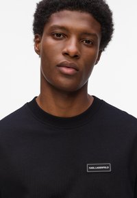 Sort sweatshirt med rund hals, ribbede ærmer og et syet mærke med "KARL LAGERFELD" i hvid på venstre bryst.