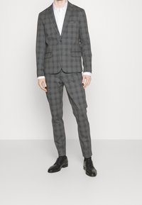 Man som bär en slim-fit grå rutiga kostym med en vit skjorta och svarta finskor, stående mot en ren vit bakgrund.