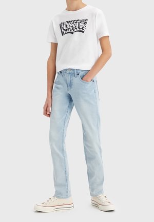 Persoon draagt een wit Levi's T-shirt met zwart geruit logo, lichtblauwe spijkerbroek en witte sneakers met zwarte en rode strepen.