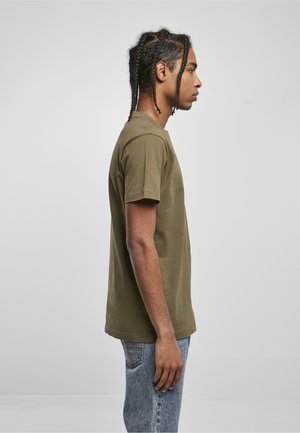 Giovane uomo con capelli intrecciati, indossa una maglietta verde oliva e jeans blu, ritratto di profilo contro uno sfondo grigio uniforme.