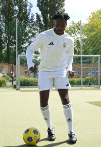 Hvid langærmet trøje med geometrisk mønster, sorte Adidas-striber, hvide shorts og sorte fodboldstøvler; spiller kontrollerer en gul/blå fodbold.