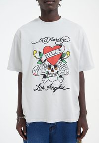 Svetle šedé tričko Ed Hardy s grafickým designem obsahujícím srdce, lebku a text "Láska zabíjí pomalu". Krátké rukávy, bavlněný materiál.