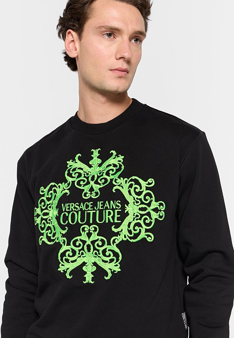 Sudadera negra con un destacado diseño bordado en verde que incluye el texto "Versace Jeans Couture" y detalles ornamentales en el centro.