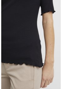 Black ribbed top with short sleeves and scalloped edges, paired with light beige pants featuring side pockets and stitched details.