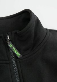 Czarna kurtka zapinana na zamek z wysokim kołnierzem, z zielonym uchwytem do zamka "HI THERE" i gładkim, strukturalnym materiałem.