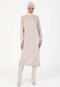 Beiges Strickkleid mit gerippter Textur, langen Ärmeln und hohem Kragen. Kombiniert mit hohen, hellen Stiefeln.