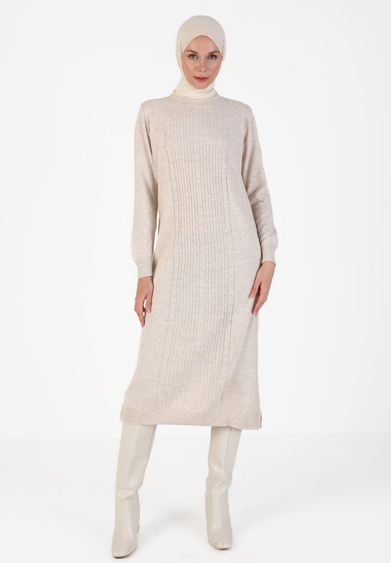 Beiges Strickkleid mit gerippter Textur, langen Ärmeln und hohem Kragen. Kombiniert mit hohen, hellen Stiefeln.