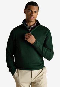 Muži, ktorý má na sebe tmavozelený sveter na krátky zips cez károvanú košeľu, s béžovými nohavicami, stojaci na bielom pozadí a drží zips blízko goliera.