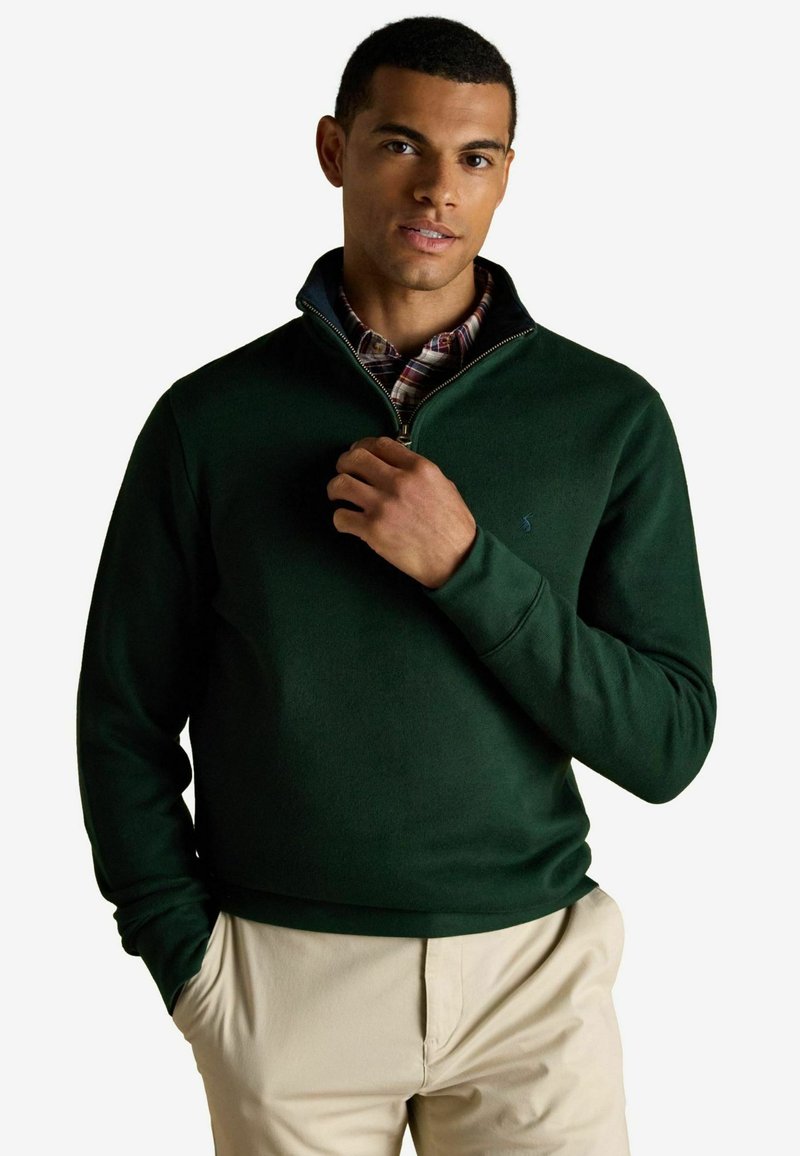 Muži, ktorý má na sebe tmavozelený sveter na krátky zips cez károvanú košeľu, s béžovými nohavicami, stojaci na bielom pozadí a drží zips blízko goliera.