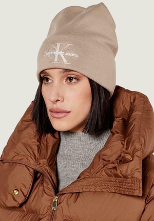 Cappello beige lavorato a maglia con logo "CK" ricamato, indossato con una giacca imbottita marrone e un dolcevita grigio, che mostra un design semplice e casual.