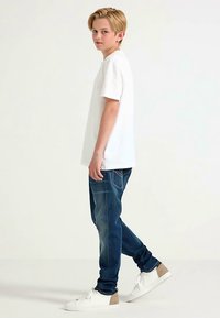 Camicia bianca a maniche corte, jeans in denim blu e sneakers bianche con accenti beige. I jeans hanno una vestibilità classica e una leggera scoloritura.