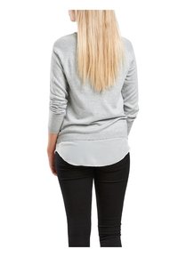Pull gris à manches longues, superposé sur une chemise gris clair, associé à un pantalon noir. Matière douce, poignets côtelés et ourlet de chemise arrondi.