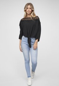 Svart bluse med blonder ved halsen og trekvart ermer, bundet foran. Brukt med lyseblå smale jeans og hvite joggesko.