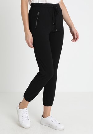 Personne portant un pantalon de jogging noir avec une poche zippée et des baskets blanches, debout devant un fond clair uni.