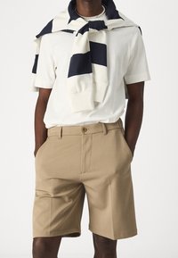 Weißes Baumwoll-T-Shirt kombiniert mit khakifarbenen Shorts in geradem Schnitt. Ein dunkelblauer und weiß gestreifter Pullover ist lässig um die Schultern gebunden.