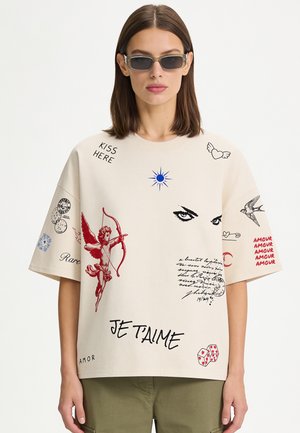 Femme aux cheveux bruns raides portant des lunettes de soleil rectangulaires et un t-shirt beige oversize avec des imprimés graphiques rouges et noirs ainsi qu'un pantalon vert.