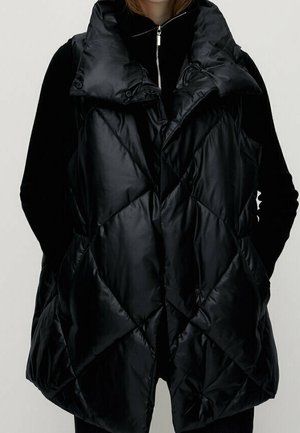 Ærmeløs sort dunvest med bred krave båret over en sort langærmet top, hænderne puttet i sidelommerne.