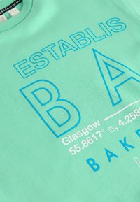 Mintgrön sweatshirt med fet blå text som läser "ESTABLIS" och "BAKED." Inkluderar Glasgow-koordinater i vit tryck. Mjuk textur.