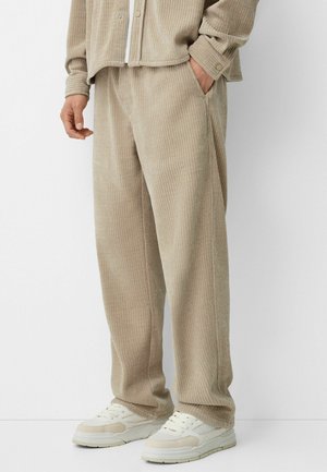 Beżowe prążkowane spodnie ze sztruksu noszone z dopasowaną kurtką i białymi trampkami, osoba stoi z jedną ręką w kieszeni.