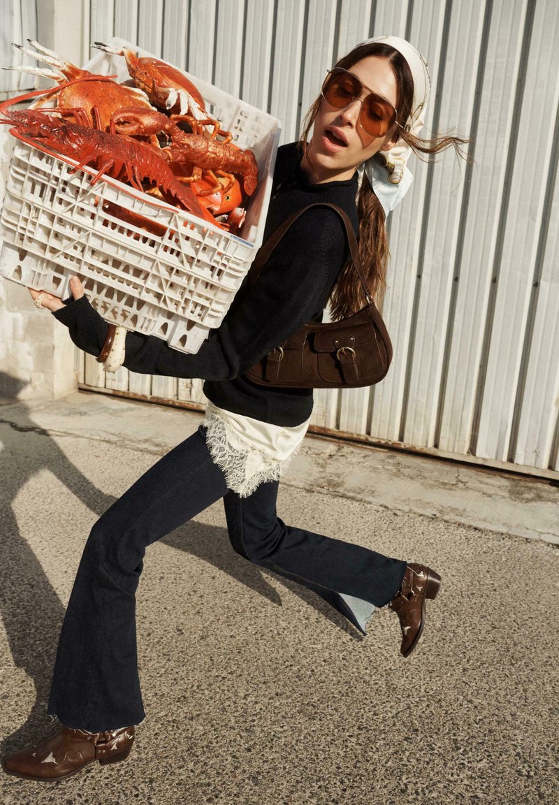 Femme portant des lunettes de soleil et un foulard, courant en plein air avec une caisse remplie de grosses langoustes cuites.