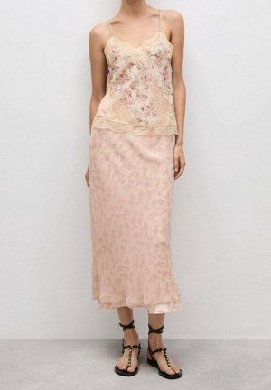 Mujer que lleva un camisón de encaje floral beige y una falda midi a juego, combinados con sandalias negras con tachuelas, de pie contra una pared blanca lisa.