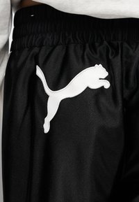 Κοντινό πλάνο μαύρου αθλητικού παντελονιού με ελαστική μέση και λευκό λογότυπο πούμα σε κίνηση στο αριστερό μηρό, πάνω σε ανοιχτό γκρι φόντο.