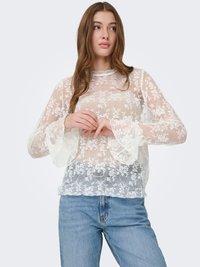 Blouse en dentelle avec broderies florales, texture transparente, manches longues évasées et ourlet festonné, associée à un jean en denim bleu clair.